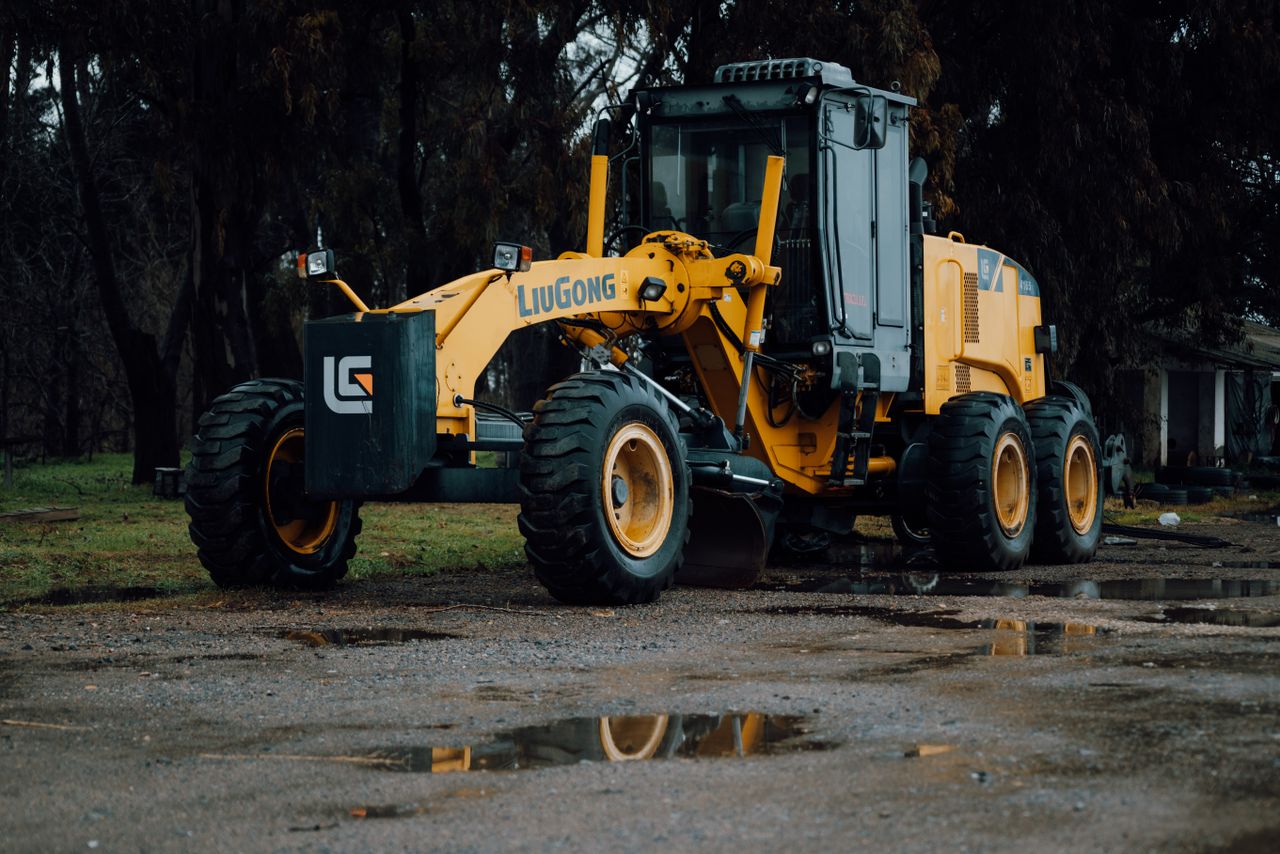 A yellow LiuGong road grader parked on wet pavement surrounded by trees.